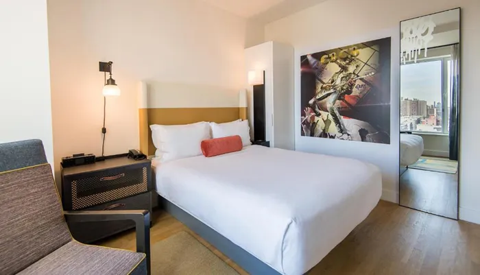 The image shows a modern hotel room with a large bed contemporary furnishings and an abstract art piece above the headboard with a view of the cityscape visible through a window