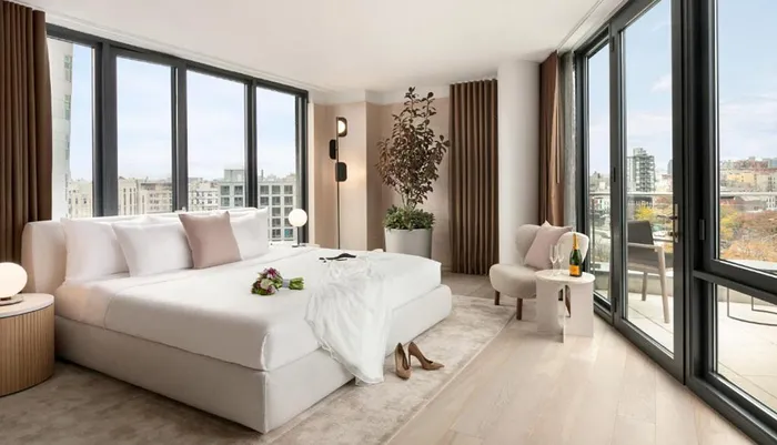 The image shows a modern and elegantly furnished bedroom with large windows offering a city view a plush king-sized bed contemporary decor and a bottle of champagne on a side table suggesting a luxurious and comfortable living space