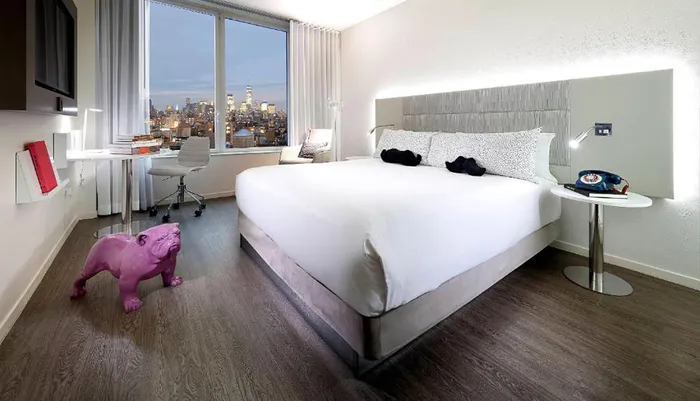 A modern hotel room with stylish decor and a striking city view through large windows featuring an unusual pink bulldog sculpture