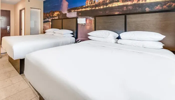 The image shows a modern hotel room with two neatly made beds and a large panoramic photograph of a scenic landscape above the headboards