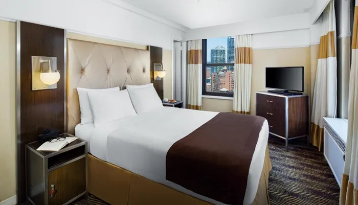The image shows a neatly arranged modern hotel room with a large bed a flat-screen TV and a window offering a view of a cityscape