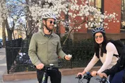 Two people wearing helmets and sunglasses are smiling while riding bicycles by a flowering tree on a sunny day.