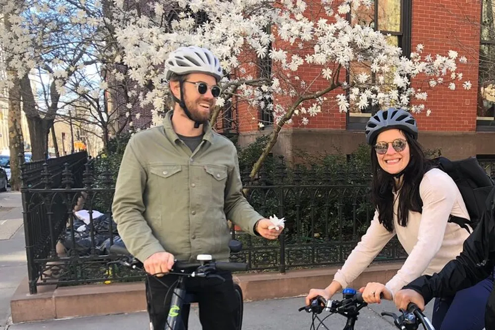 Two people wearing helmets and sunglasses are smiling while riding bicycles by a flowering tree on a sunny day