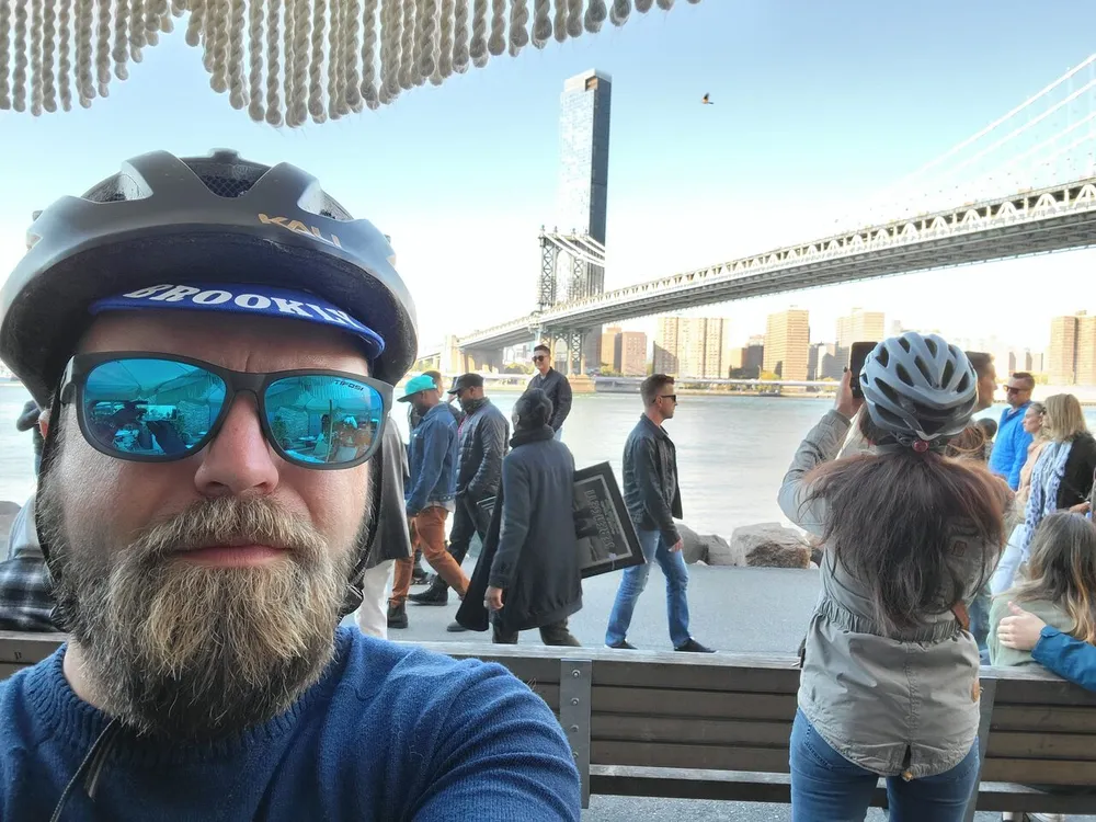 A man wearing a bicycle helmet and sunglasses takes a selfie with a bridge and people in the background
