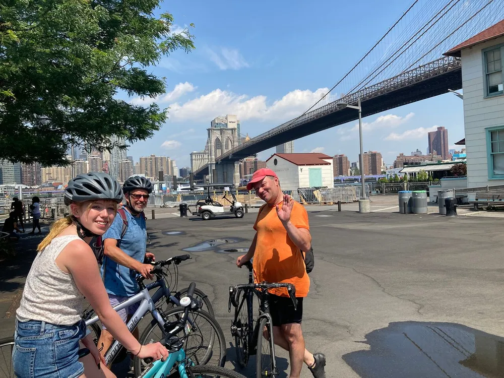 Three cyclists are smiling and posing for the camera with the Brooklyn Bridge in the background on a sunny day