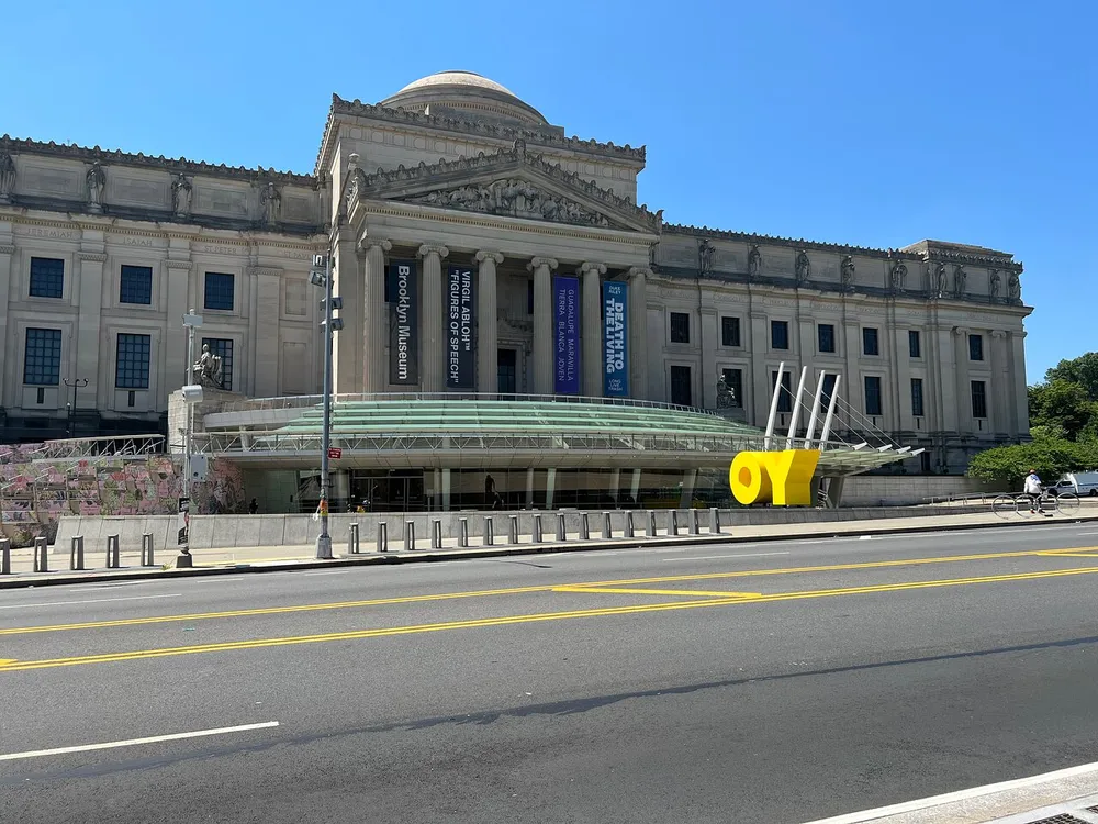 The image shows the exterior of the Brooklyn Museum in New York City featuring an OY sculpture on the sidewalk