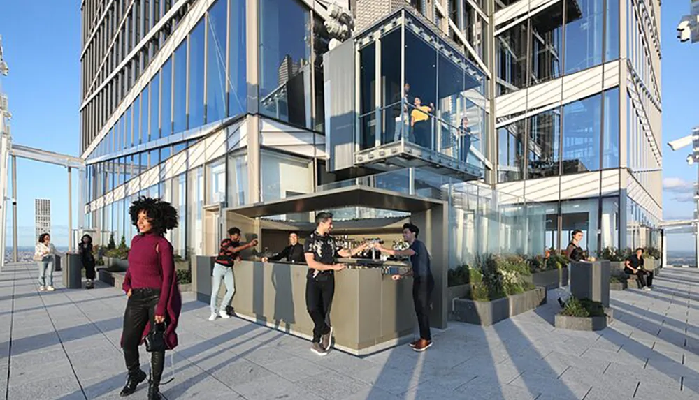 The image shows people enjoying their time in an urban rooftop setting with a bar area and a glass-enclosed balcony overlooking the city