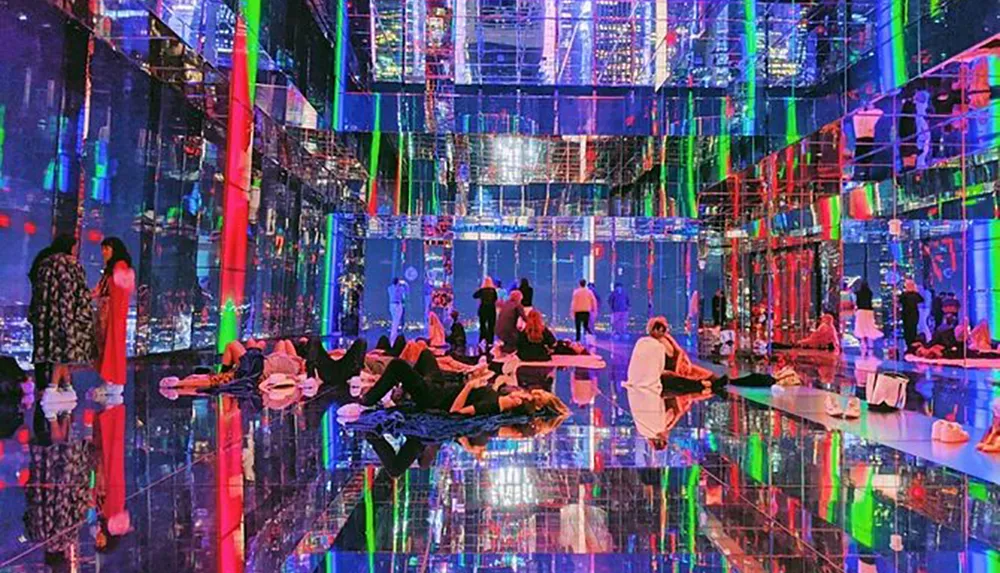 The image shows a vibrant colorful mirrored room filled with people walking and lounging creating multiplied reflections in a visually immersive space