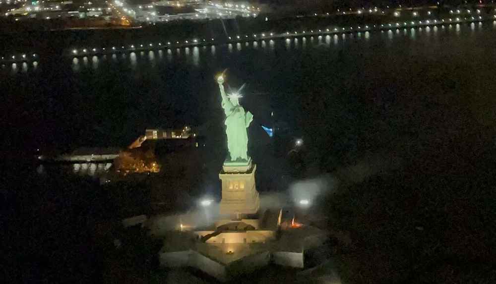 The image shows an aerial nighttime view of the Statue of Liberty illuminated against the dark surroundings with a backdrop of city lights in the distance