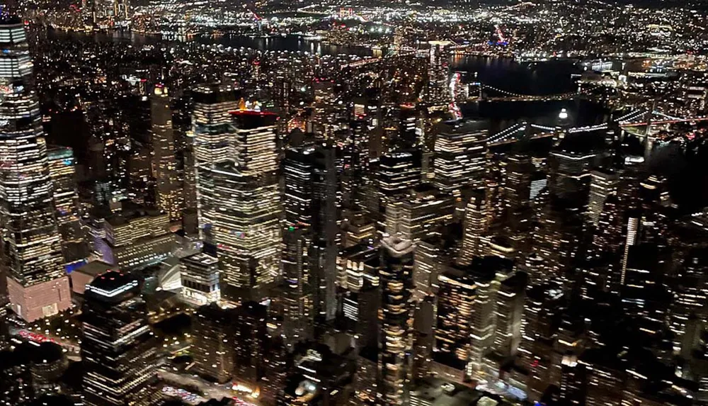 This image captures a stunning nighttime aerial view of a densely populated cityscape with illuminated skyscrapers and several bridges spanning across a river