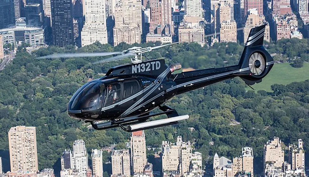 A black helicopter is flying over a city with a large park and dense skyscrapers in the background
