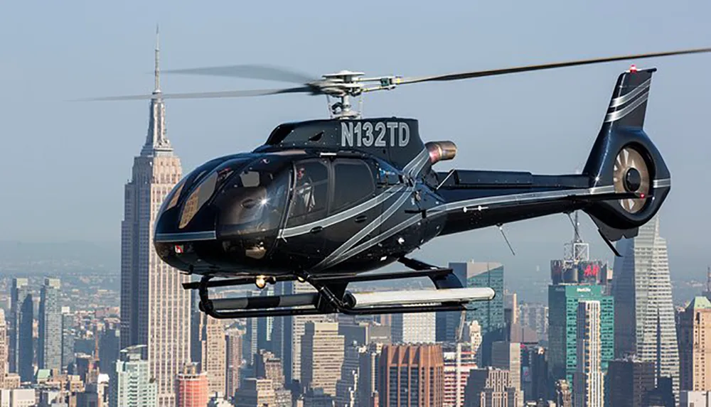 A black helicopter with registration N132TD is flying in front of a city skyline featuring the Empire State Building