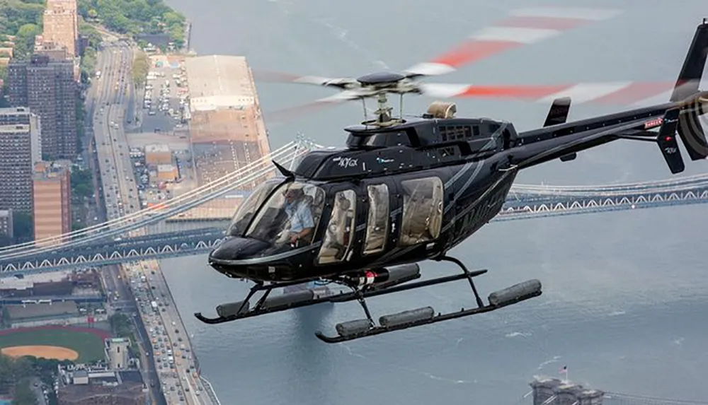A black helicopter is flying over a bridge and a baseball field with a cityscape in the background