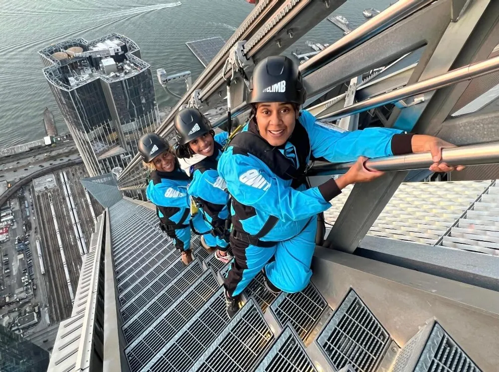 Three individuals in blue jumpsuits and helmets are perched on the edge of a high structure above a cityscape engaging in an urban outdoor adventure