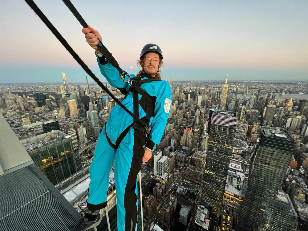 A person in a blue jumpsuit with safety harnesses is standing on the edge of a skyscraper with the New York City skyline in the background during twilight