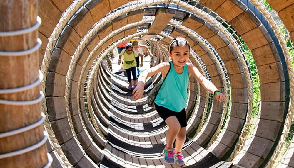 A young girl is leading the way through a cylindrical wooden playground tunnel with people following behind her