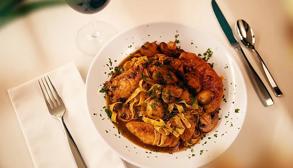 A plate of pasta with chicken and mushrooms is served on a table beside a set of silverware and a half-filled glass of red wine evoking an elegant dining experience