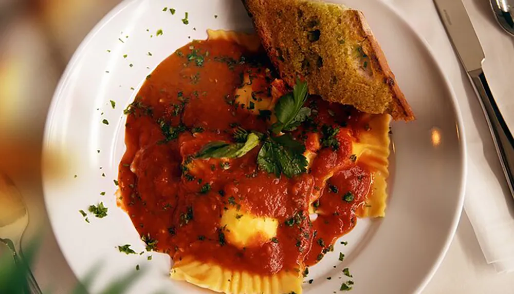 The image displays a plate of ravioli covered in tomato sauce and sprinkled with herbs accompanied by a slice of toasted garlic bread