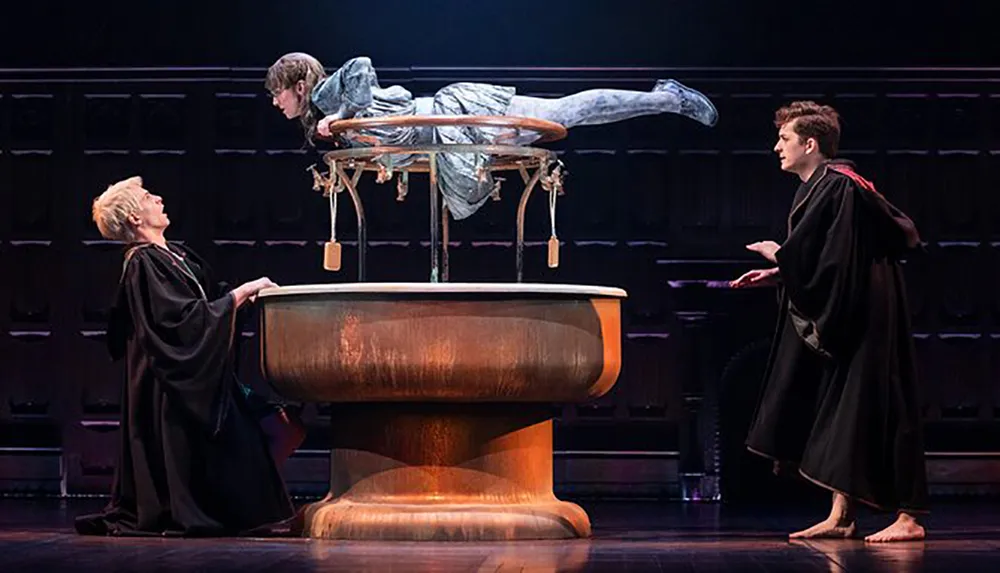 Two actors in robes perform on stage in a magical scene with a person levitating above a table as part of a theatrical production