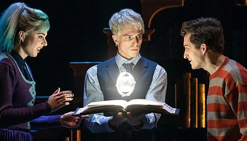 Three actors on stage perform a scene where a glowing book is being held by a man with an intrigued expression flanked by a woman with blue hair and a man in a striped sweater looking on intently