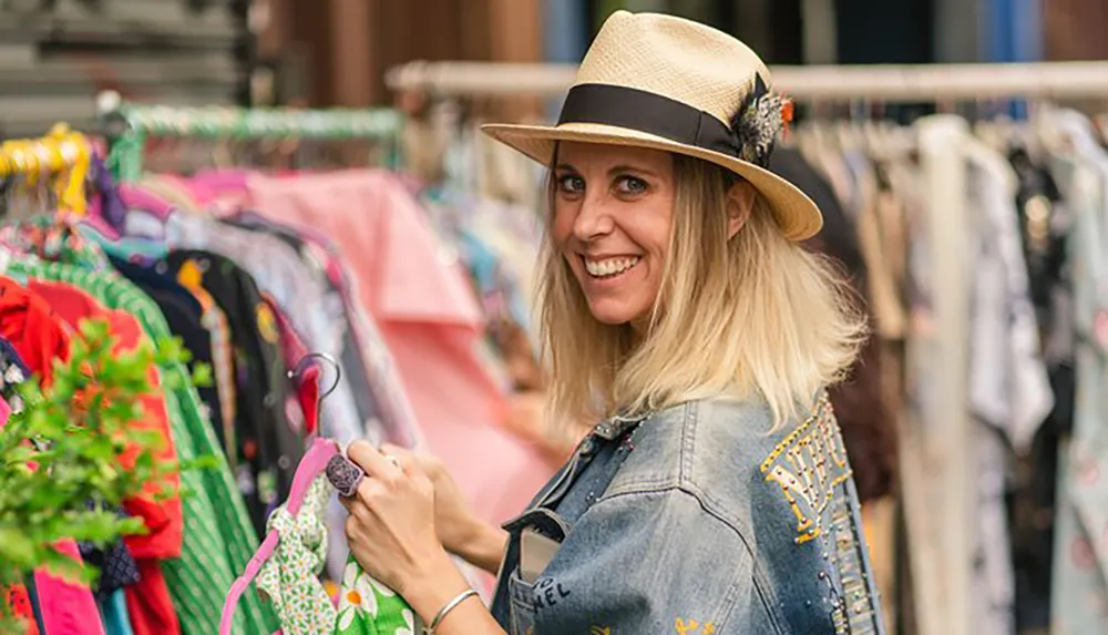 A smiling woman in a straw hat and denim jacket is browsing through clothes at an outdoor market