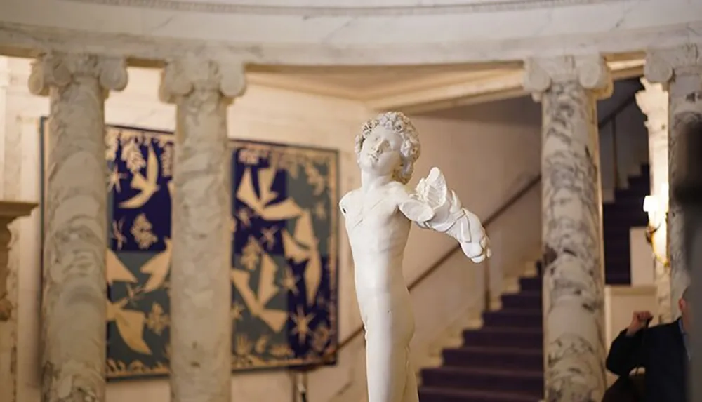 A marble statue of a cherubic figure extends its arm in a grand classical interior with decorative columns and tapestries