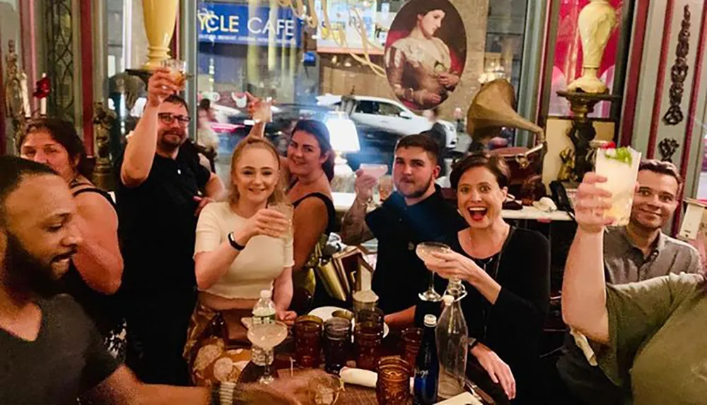 A group of people is joyfully toasting with drinks in a lively indoor setting with a glimpse of the city street visible through the window