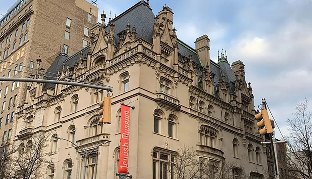 The image shows a grand Gothic Revival style building adorned with a red banner indicating it is the Jewish Museum set against a partly cloudy sky with street traffic lights in the foreground