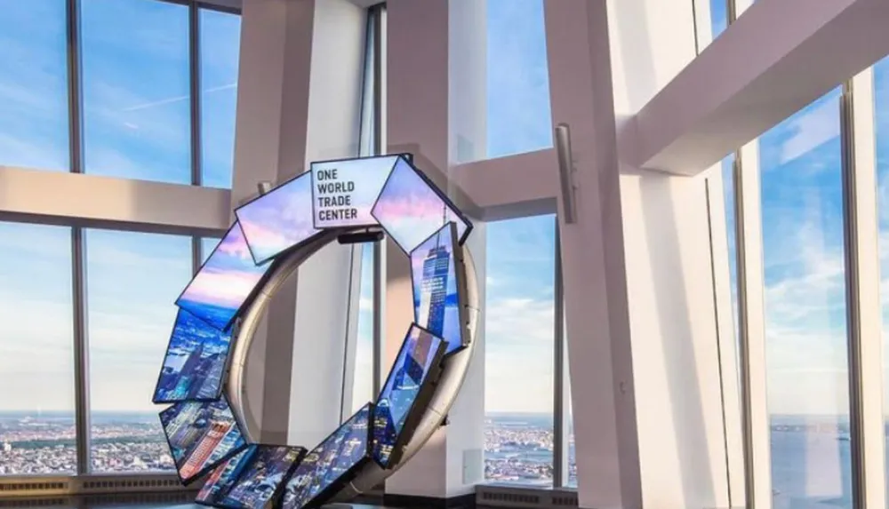 The image shows an artistic ring-shaped digital display with multiple screens labeled One World Trade Center set against a backdrop of large windows with a view of the sky and horizon