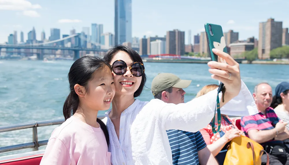 Two people are taking a selfie with a smartphone against the backdrop of a city skyline and bridge likely from a boat or waterfront