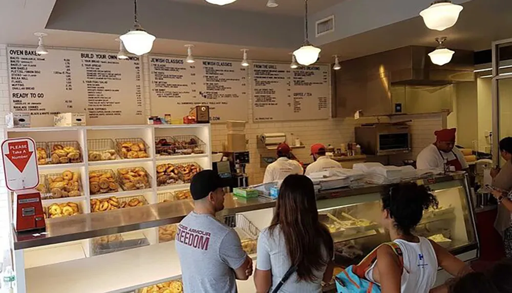 Customers are waiting in line at a deli counter with an array of baked goods on display and a menu board listing various sandwiches and dishes above