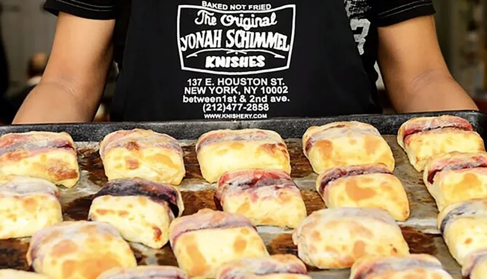 A person is presenting a tray of baked knishes in front of a sign that advertises The Original Yonah Schimmel Knishes in New York City