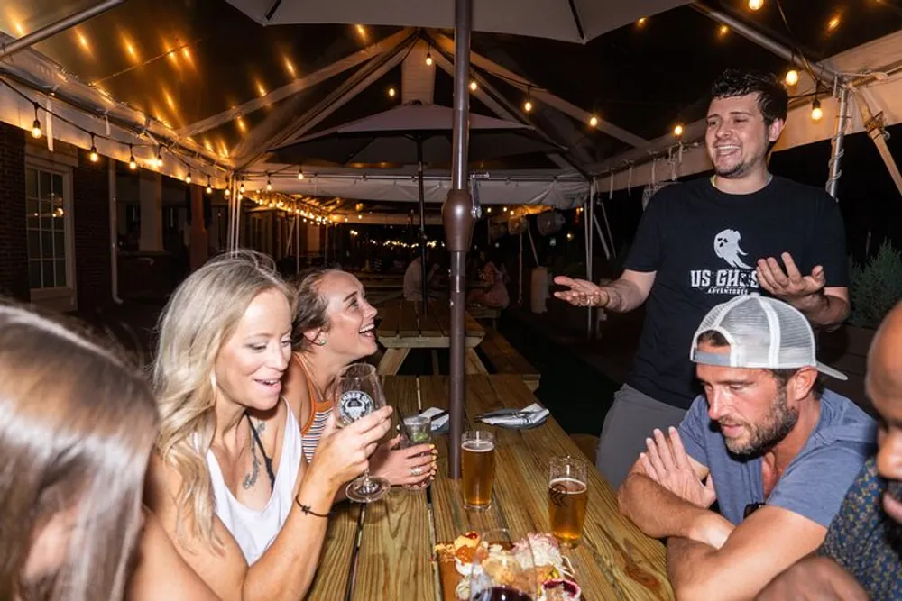A group of people is enjoying an evening at an outdoor patio socializing and laughing under string lights