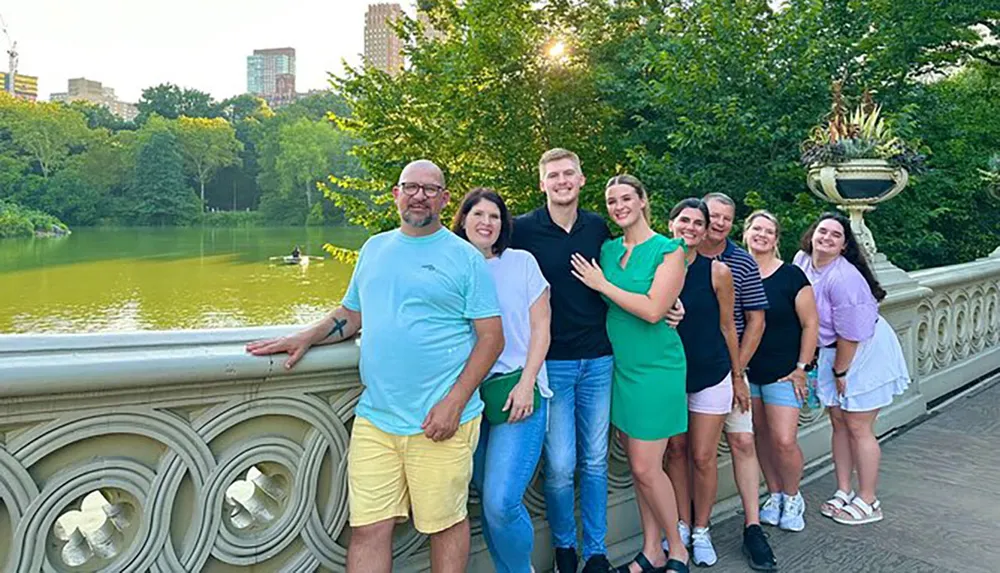 A group of eight people is posing for a smiling photo on a bridge over a pond in a lush park environment with city buildings in the background