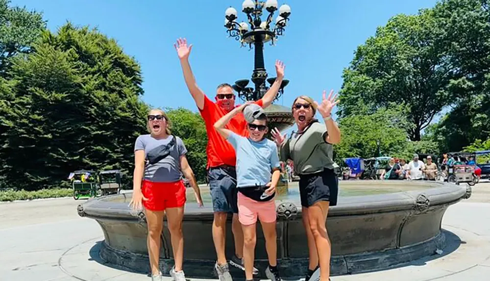 A joyful group of four is posing with raised arms and cheerful expressions in front of an ornate lamp post and fountain seemingly enjoying a sunny day outdoors