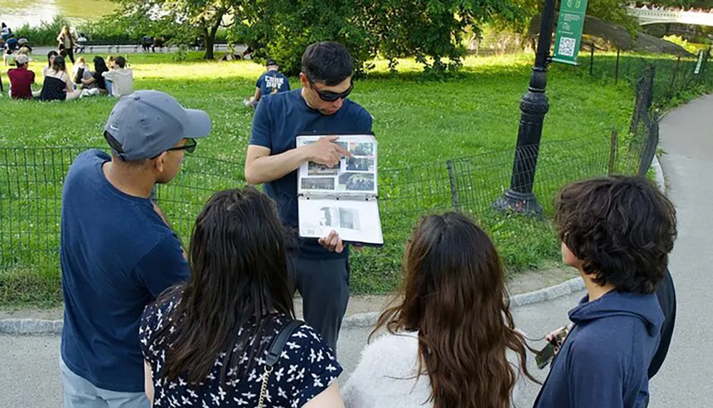 A person is showing and explaining something in a binder to a group of attentive onlookers in a park setting