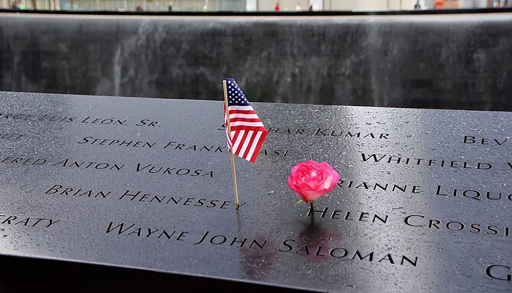 The image shows a wet dark stone memorial with engraved names adorned with an American flag and a pink rose which appears to be a tribute at a solemn site