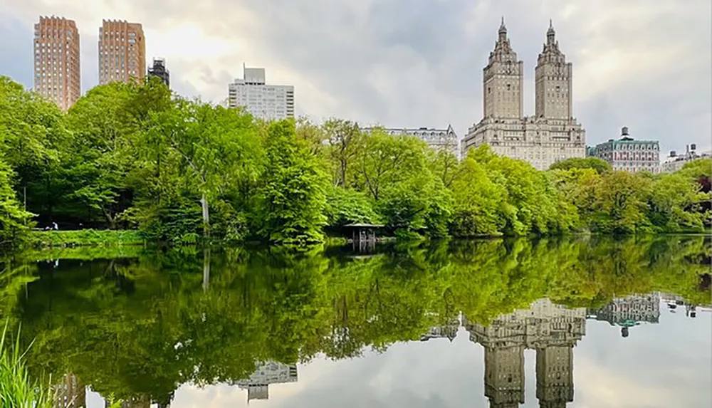 The image shows a tranquil scene in a lush park with a pond reflecting the surrounding trees and the towering architecture of adjacent buildings
