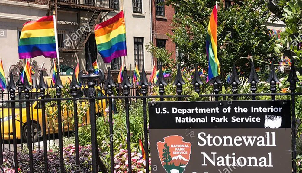 The image shows rainbow flags prominently displayed on a fence in front of a sign for the Stonewall National Monument symbolizing LGBTQ pride and history