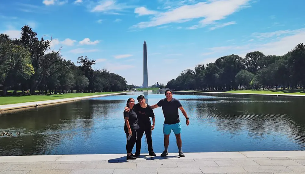 Three people are posing for a photo with the Washington Monument in the background alongside the Reflecting Pool in Washington DC