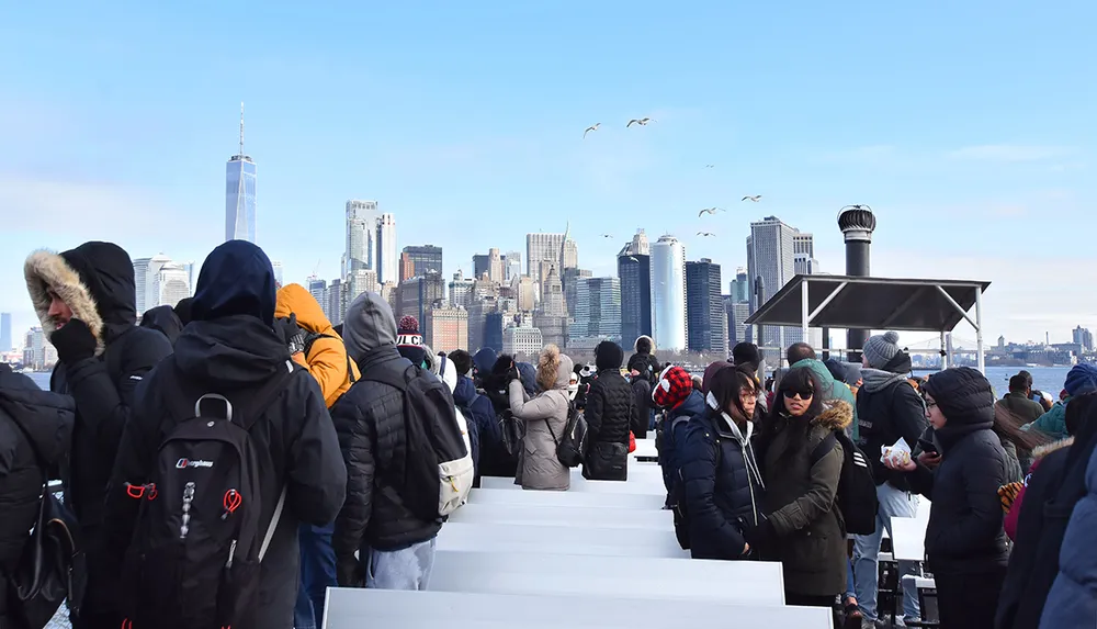 A group of people is standing on a ferry or outdoor platform in cold weather facing a scenic view of a city skyline with birds flying overhead