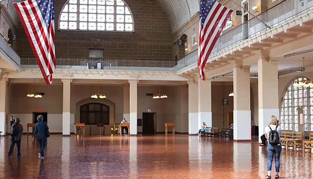 The image shows a spacious historical interior with large American flags hanging from the ceiling and visitors walking around and sitting at benches in what appears to be a public building with a grand architecture