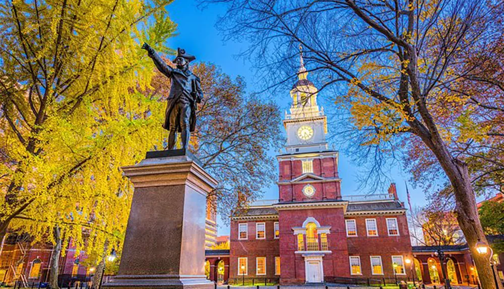 The image shows an illuminated statue in the foreground with an historic brick building adorned by an iconic clock tower in the background set among autumn-colored trees in the twilight