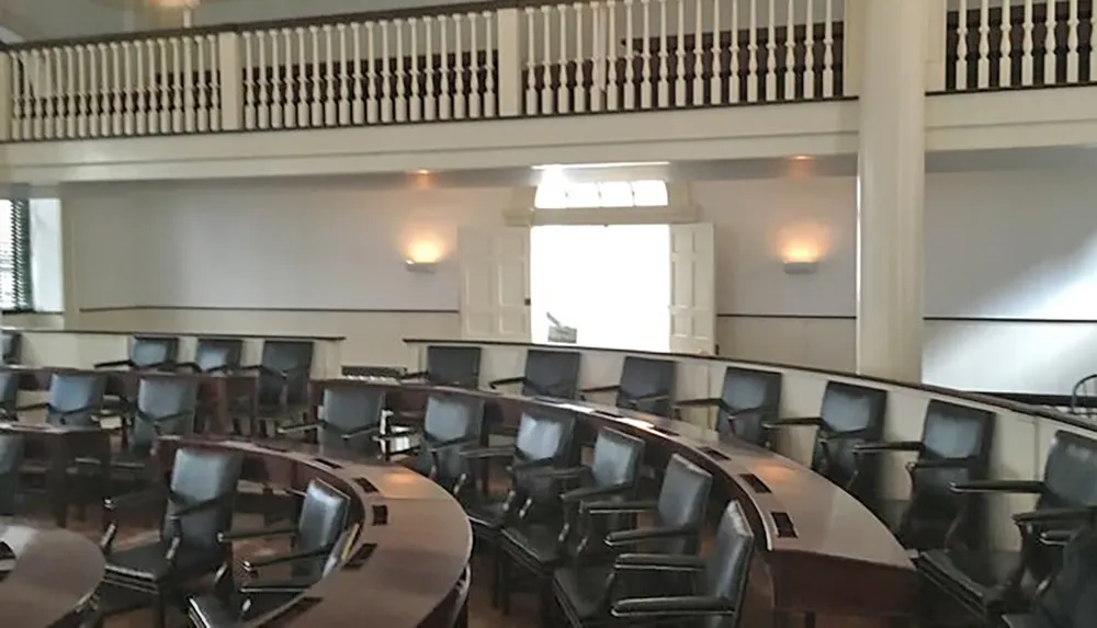 The image shows a formal meeting room with rows of upholstered chairs arranged in a semicircular pattern facing a central area with a balcony level above