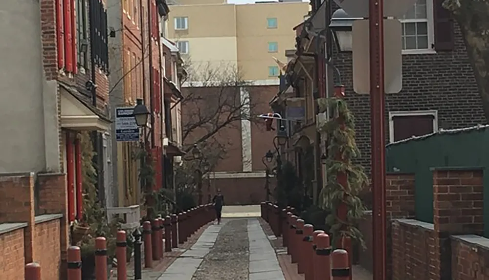 The image shows a quaint narrow alley lined with red bollards and historical buildings with a person in the distance walking away from the viewer