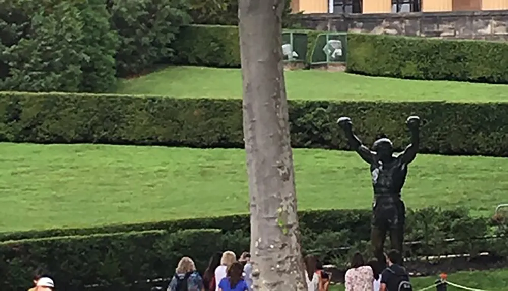 The image shows a group of people walking in a garden with well-manicured hedges and a statue featuring a figure with arms raised triumphantly