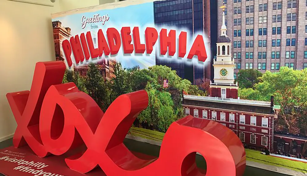 The image shows a red XOXO sculpture in the foreground with a backdrop featuring a large poster that says Greetings from Philadelphia displaying iconic architecture of the city