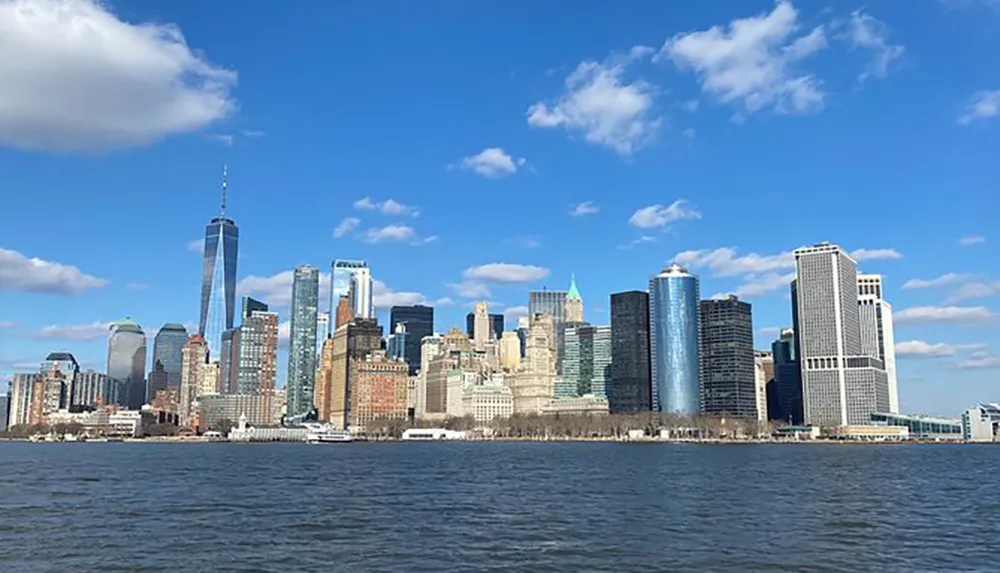 The image shows a clear view of a city skyline with skyscrapers standing tall under a blue sky interspersed with clouds as seen across a stretch of water
