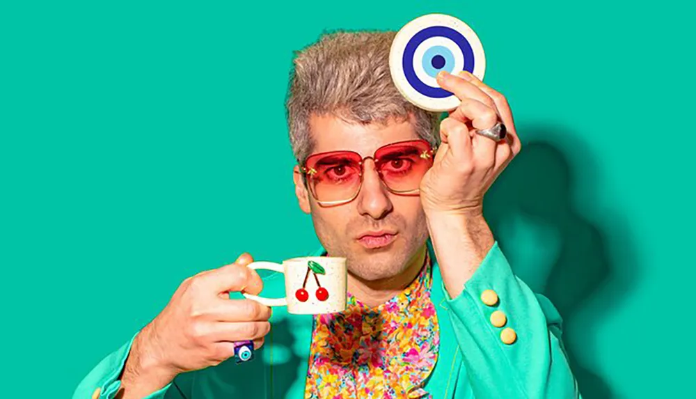 A man with silver hair wearing red-tinted sunglasses and a colorful floral shirt under a teal blazer holds a whimsical teacup in one hand and balances a target on the tip of his finger in front of a turquoise background