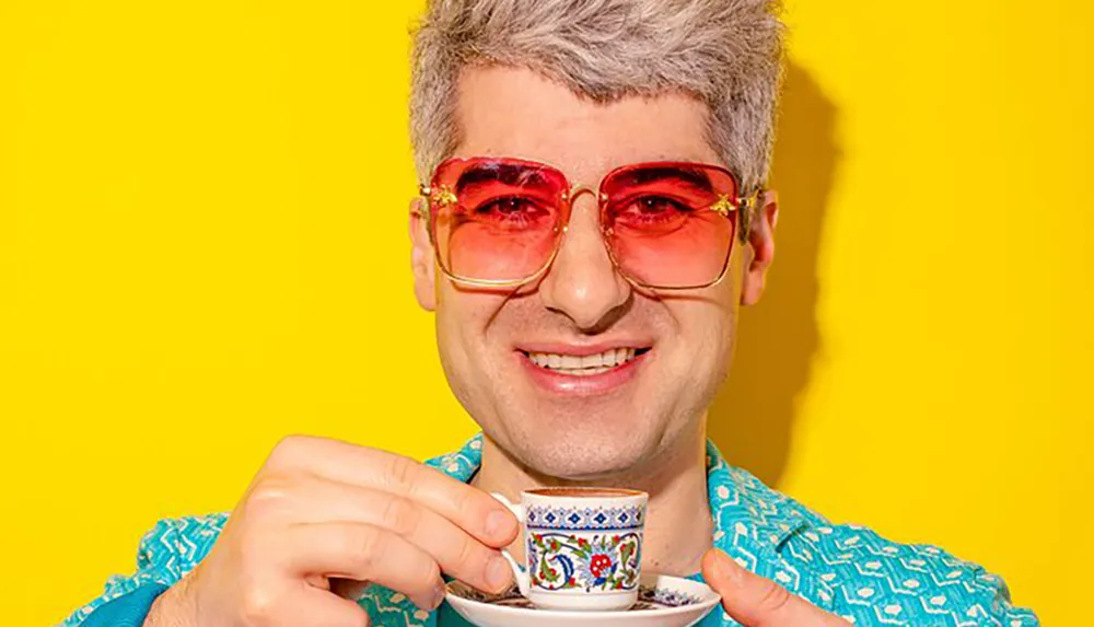 A person with silver hair and red-tinted glasses smiles at the camera while holding a decorative cup and saucer against a bright yellow background